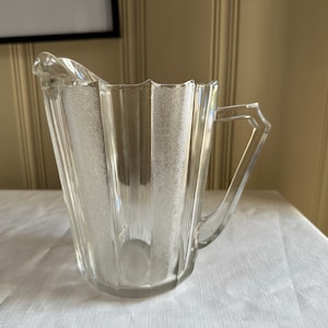 May include: A clear glass pitcher with a textured, frosted design. The pitcher has a handle and a spout, and it sits on a white tablecloth. The pitcher is likely used for serving beverages.
