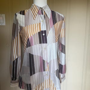 May include: A long-sleeved button-down shirt with a geometric pattern in shades of brown, white, and purple. The shirt is displayed on a mannequin and features a collared neckline and buttoned cuffs. The design includes vertical and angled stripes.
