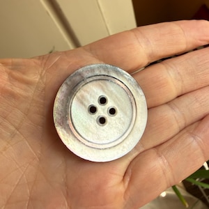 May include: A round, iridescent mother-of-pearl button featuring a smooth, polished surface with a subtle sheen. The button has a recessed center area containing four evenly spaced holes for attachment, surrounded by a raised, circular outer rim.