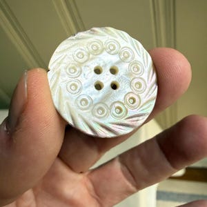 May include: A round, iridescent mother-of-pearl button featuring a central four-hole design surrounded by a ring of small, circular indentations. The outer edge of the button is decorated with a series of diagonal, carved grooves.