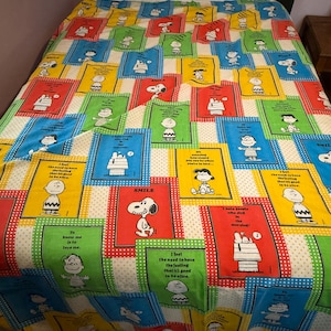 May include: A colorful bedspread featuring a repeating pattern of Snoopy and other Peanuts characters. The design includes various colored squares with quotes and the word "SMILE". The bedspread has a white background with a dotted border.
