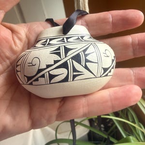 May include: A small, cream-colored ceramic ornament shaped like a miniature pot, featuring intricate black geometric patterns and lines. A thin black ribbon is attached to the top, allowing it to be hung as a decorative piece for display.