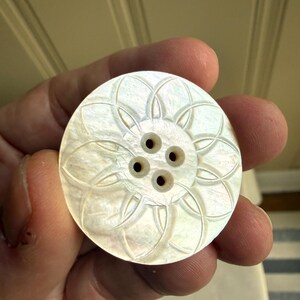 May include: A round, iridescent mother-of-pearl button featuring a carved, symmetrical floral pattern radiating from the center. The button has four small, evenly spaced holes in the middle for attachment, with a smooth, polished surface finish.