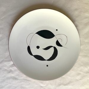 May include: White ceramic plate with a black abstract design. The plate has a thin red rim and a minimalist, artistic pattern of curved shapes and dots. The design is centered on the plate.