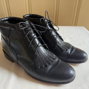May include: A pair of black leather ankle boots with a textured finish. The boots feature a lace-up design with decorative fringe on the front. The boots are displayed on a white surface.