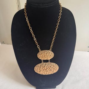 May include: A gold-toned necklace featuring a thick chain and two stacked, oval-shaped pendants. Both pendants have a textured, hammered surface with raised bumps. The smaller oval sits above the larger oval, and both are connected by small metal rings.