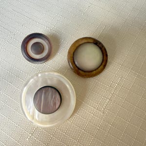 May include: Three circular buttons of varying sizes. The smallest button is dark purple with a lighter center. The medium button has a brown outer ring and a cream-colored center. The largest button features a white pearlescent rim and a dark center.