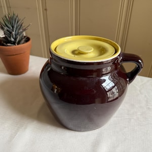 May include: A brown ceramic crock with a yellow lid and a handle. The crock is glazed and has a smooth finish.