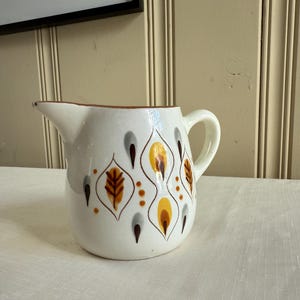 May include: White ceramic pitcher with a brown rim and handle. The pitcher is decorated with a repeating pattern of stylized leaves in brown, yellow, and gray. The pitcher has a spout for pouring.