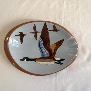 May include: Oval ceramic ashtray with a brown rim and a light blue interior. The ashtray features a painted design of a flying goose with outstretched wings and three smaller geese in flight. The geese are brown, white, and black.