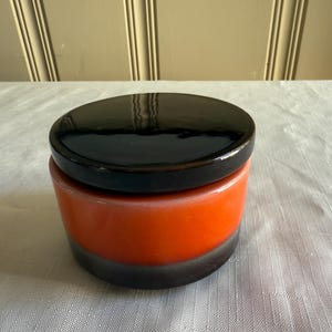 May include: A ceramic jar with a glossy black lid and a vibrant orange body. The base of the jar is a dark gray color. The jar is sitting on a white surface.