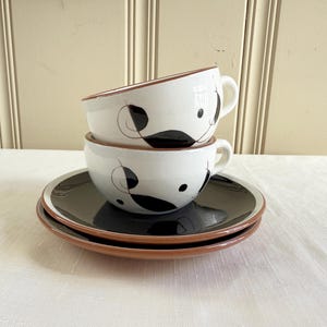 May include: A stack of two white ceramic coffee cups with black abstract designs and brown rims, resting on two matching black plates with brown rims. The cups have handles.