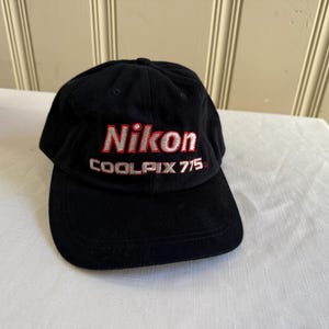 May include: Black baseball cap with the Nikon logo embroidered in red and white. The text "COOLPIX 775" is also embroidered on the cap. The cap is sitting on a white surface.