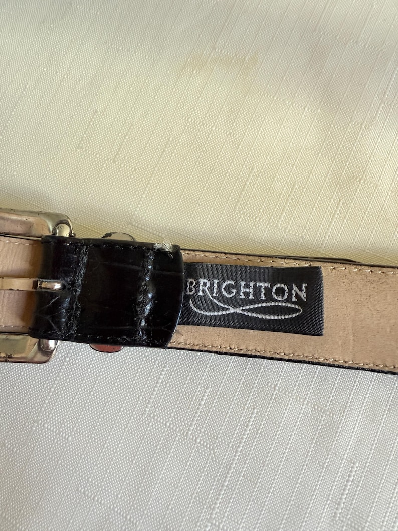 Może przedstawiać: Czarny sk&oacute;rzany pasek ze srebrną klamrą. Pasek ma teksturowany wz&oacute;r przypominający krokodyla. Nazwa marki "BRIGHTON" jest wyświetlana na czarnej etykiecie.