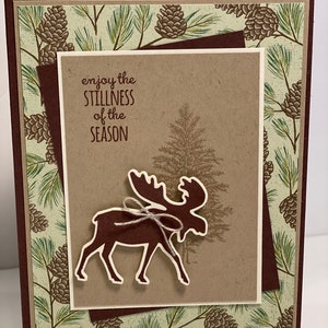 May include: A handmade greeting card with a brown moose silhouette and the text "enjoy the stillness of the season." The card features a layered design with a pine cone and pine needle pattern in shades of green and brown.