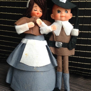 May include: Two vintage Thanksgiving dolls. One doll is a woman in a blue dress, white apron, and brown cape. The other doll is a man in a brown outfit and black hat. Both dolls have painted faces.