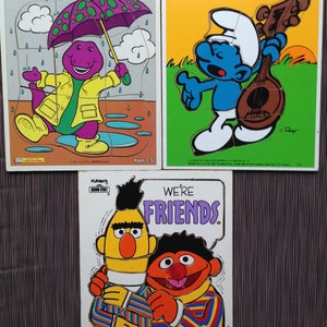May include: Three vintage Playskool wooden jigsaw puzzles. One features Barney the dinosaur in a yellow raincoat and purple umbrella. Another shows a Smurf playing a mandolin. The third puzzle says "We're Friends" with Bert and Ernie.