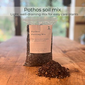 May include: A bag of Pothos potting mix on a wooden table. The bag is labelled "Pothos Potting Mix" and "Well-draining blend designed for healthy pothos plants." A small pile of the mix is next to the bag. The text "Pothos soil mix" is at the top.