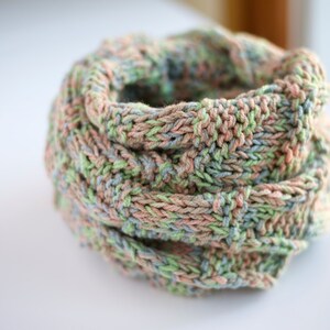May include: A soft, knitted scarf in shades of pink, green, and blue. The scarf is folded and rolled into a circle.