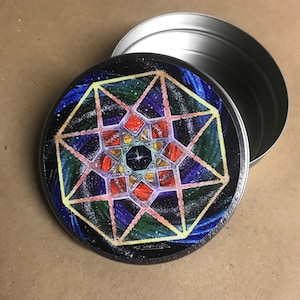 May include: A round metal tin with a colorful geometric design on the lid. The design features a starburst pattern with shades of blue, green, red, yellow, and orange. The tin is open and the inside is visible.