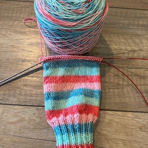 Può includere: Un gomitolo di filato colorato nei toni del blu, rosa e verde acqua è accanto a una calza parzialmente lavorata a maglia. La calza presenta strisce orizzontali degli stessi colori, con un polsino a coste e un motivo strutturato sul piede. Ferri da maglia inseriti.