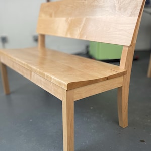 May include: A light-colored wooden bench with a backrest. The bench has a rectangular seat and four sturdy legs. The backrest is angled and provides support. The wood grain is visible, and the bench appears to be newly constructed.