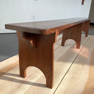 Peut inclure: Banc en bois artisanal avec une finition marron foncé. Le banc a un dessus rectangulaire aux bords arrondis et des pieds arqués. Le grain du bois est visible, mettant en valeur la beauté naturelle du matériau. Le banc mesure environ 150 cm de long.