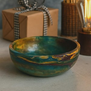 Può includere: Una ciotola di legno fatta a mano con una vibrante combinazione di colori a strati di verde acqua, giallo e marrone. La ciotola è rotonda e poggia su una superficie chiara. Una confezione regalo avvolta e elementi decorativi sono sullo sfondo.