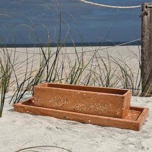 Puede incluir: Una jardinera rectangular de color terracota con una base a juego se encuentra en una playa de arena. La escena incluye una valla de cuerda, postes de madera y hierba de playa, con un cielo nublado en el fondo. La jardinera mide aproximadamente 46 cm de largo.