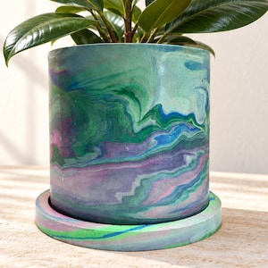 May include: A cylindrical plant pot with a matching circular saucer, featuring a marbled pattern of swirling green, blue, purple, and pink colours. A green-leafed rubber plant with a single reddish-brown new leaf grows from the pot.