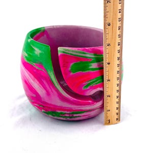 Handmade yarn bowl with bright pink, green, and white marbled swirl design, shown with a ruler for scale to display its compact size, perfect for holding standard yarn cakes and skeins. This smooth, glossy yarn holder features a curved yarn guide slot to prevent tangles while knitting or crocheting. A colorful knitting bowl ideal for crochet projects, fiber arts, craft rooms, and thoughtful gifts for knitters, crocheters, and yarn lovers who appreciate unique handmade tools.