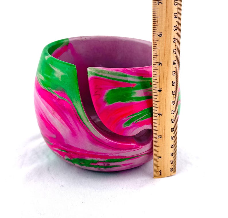 Handmade yarn bowl with bright pink, green, and white marbled swirl design, shown with a ruler for scale to display its compact size, perfect for holding standard yarn cakes and skeins. This smooth, glossy yarn holder features a curved yarn guide slot to prevent tangles while knitting or crocheting. A colorful knitting bowl ideal for crochet projects, fiber arts, craft rooms, and thoughtful gifts for knitters, crocheters, and yarn lovers who appreciate unique handmade tools.