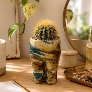 May include: A unique, handcrafted ceramic planter in a marbled pattern of blue, brown, and beige. The planter is shaped like a pair of legs and holds a small, spiky cactus. The cactus is green with yellow spines.