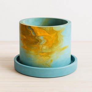 May include: A cylindrical teal and gold marbled planter with a matching teal saucer. The planter has a smooth, glossy finish and is designed to hold small plants or succulents. The planter is approximately 5 inches tall and 5 inches in diameter.