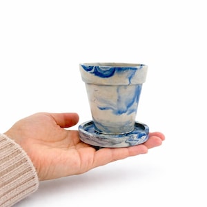 May include: A small, tapered plant pot with a matching circular saucer, featuring a marbled pattern of white and blue swirls. The surface has a smooth, matte finish with distinct blue pigment streaks integrated into the light-colored base material.