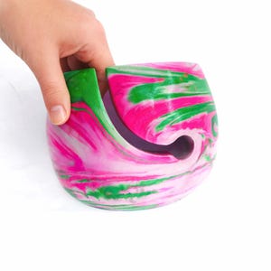 Bright pink and green marbled cement yarn bowl shown in hand, highlighting its smooth, sturdy weight and glossy finish. Concrete yarn holder for knitters and crocheters features a curved cutout to guide the strand, keeping yarn balls and cakes from tangling or rolling away, a colorful handmade knitting accessory and unique gift for fiber artists who love bold, modern craft room decor.