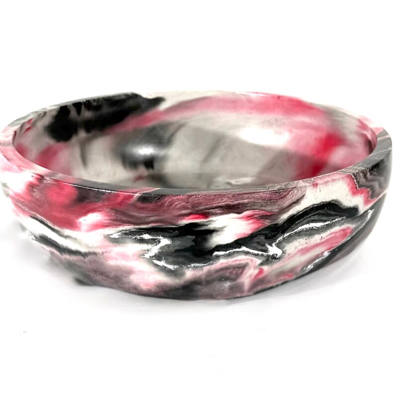 May include: A round, shallow bowl featuring a marbled pattern with swirling colors of black, white, and vibrant pink. The smooth, polished surface displays a continuous, fluid design that wraps around the exterior and interior of the vessel.