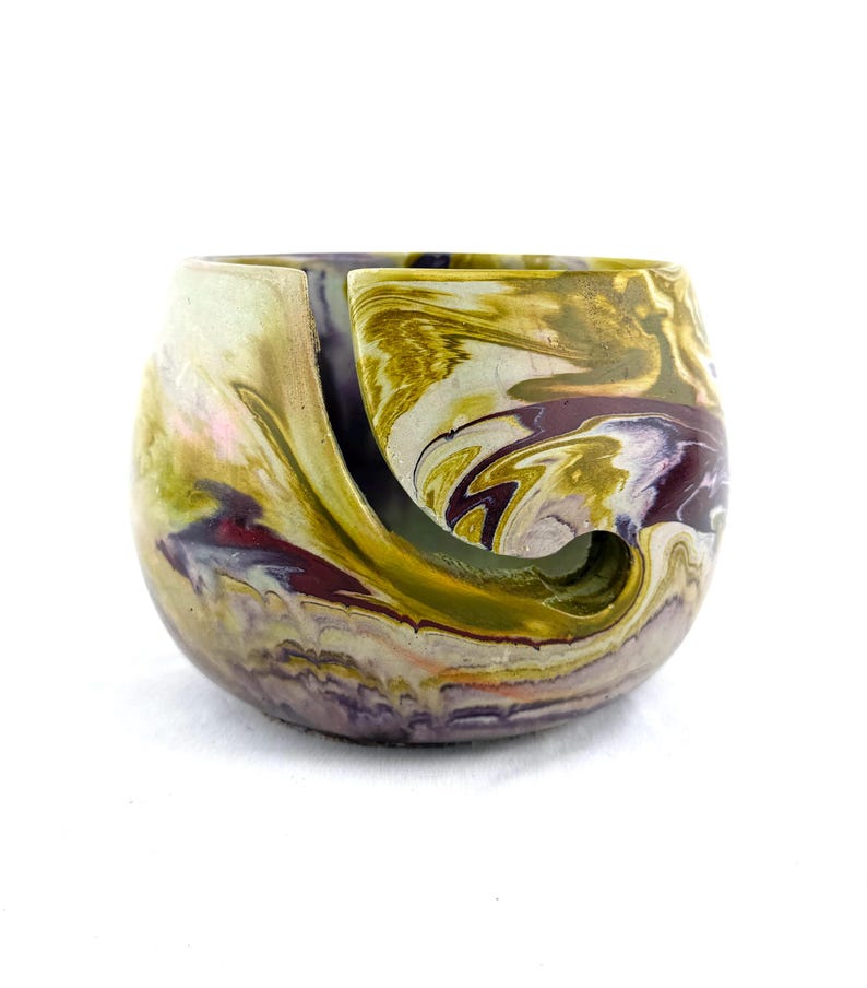 Handmade marbled cement yarn bowl in earthy mustard, plum, and cream tones with a smooth spiral cutout to guide the strand. Modern concrete yarn holder for knitters and crocheters keeps yarn cakes and balls from tangling or rolling, a durable artisan knitting accessory and unique gift for fiber artists who love colorful, contemporary craft room decor.