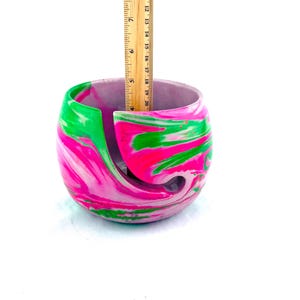 May include: A marbled yarn bowl in shades of pink, green, and white. The bowl has a curved design with a slot to feed yarn through. The bowl is approximately 6 inches in diameter and 4 inches tall.