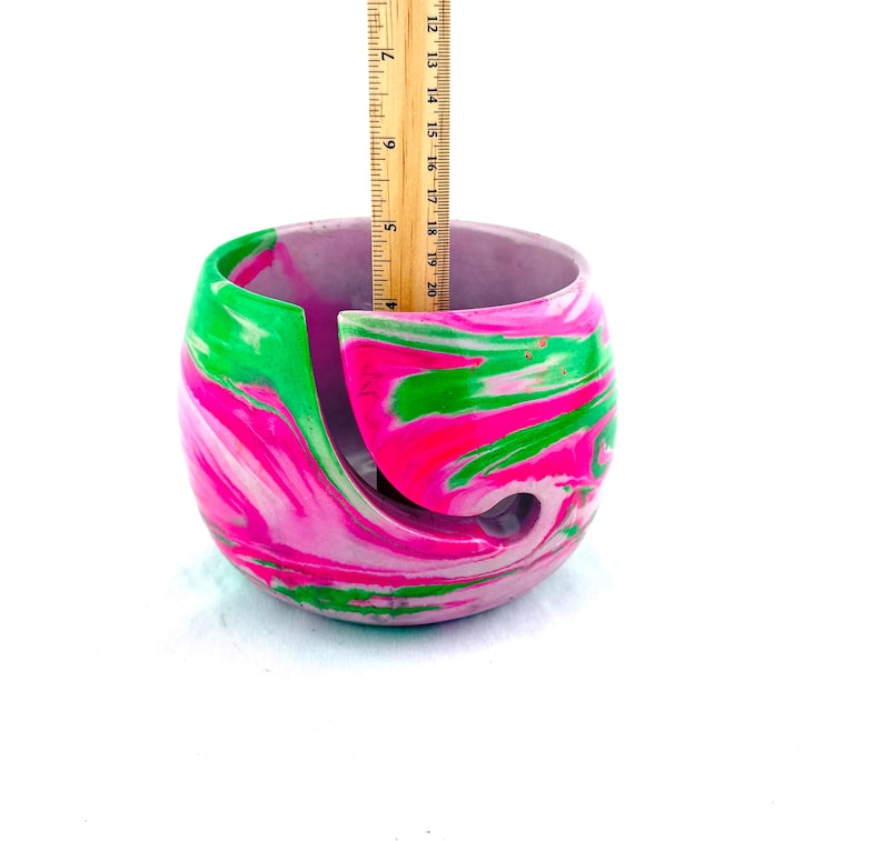 May include: A marbled yarn bowl in shades of pink, green, and white. The bowl has a curved design with a slot to feed yarn through. The bowl is approximately 6 inches in diameter and 4 inches tall.