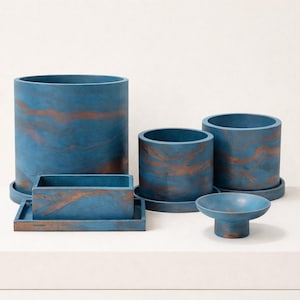 May include: A collection of blue and copper-toned concrete planters and containers. The set includes cylindrical pots of varying heights, a rectangular tray, and a small bowl. The color scheme features a marbled effect, adding visual interest to the modern design.