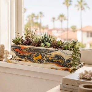 May include: A rectangular planter with a marbled pattern in shades of black, white, orange, and yellow, featuring a matching tray. The planter is filled with a variety of green and purple succulents, including a trailing string of pearls plant.