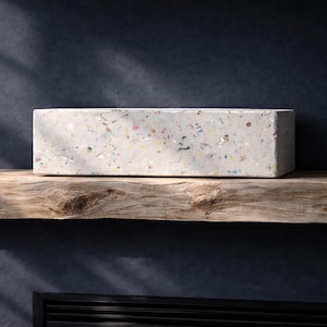 Op de afbeelding: Een rechthoekig, lichtgrijs decoratief blok met kleurrijke vlekken rust op een rustieke houten plank. Het blok staat tegen een donkerblauwe muur, met schaduwen van zonlicht die een dramatisch effect creëren. De plank heeft een natuurlijke, verweerde uitstraling.
