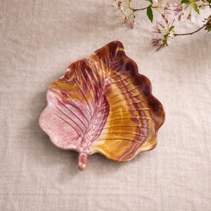 Puede incluir: Un plato de cerámica con forma de hoja y superficie texturizada. El plato presenta una mezcla de colores, incluyendo tonos de rosa, amarillo y marrón. El diseño de la hoja tiene un tallo y vetas detalladas. El plato se utiliza probablemente para servir o decorar.