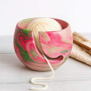Pink and green marbled cement yarn bowl holding a cream ball of yarn, with the strand feeding smoothly through a curved cutout. Modern concrete yarn holder for knitters and crocheters keeps yarn from tangling or rolling away perfect gift for fiber artists who love colorful beachy craft room decor and durable handmade knitting accessories. available in customized colors to fit your style and gift needs. packed in recycled shredded paper for eco friendly gift giving for her.