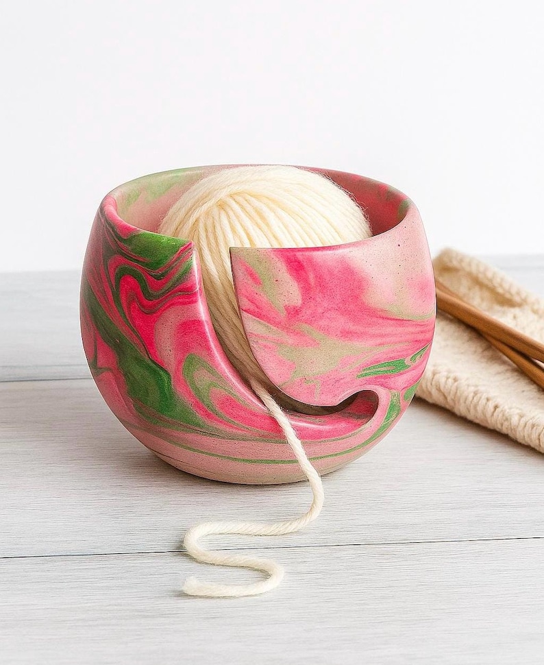 Pink and green marbled cement yarn bowl holding a cream ball of yarn, with the strand feeding smoothly through a curved cutout. Modern concrete yarn holder for knitters and crocheters keeps yarn from tangling or rolling away perfect gift for fiber artists who love colorful beachy craft room decor and durable handmade knitting accessories. available in customized colors to fit your style and gift needs. packed in recycled shredded paper for eco friendly gift giving for her.