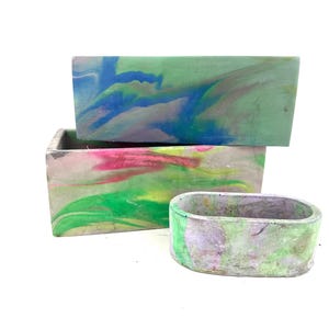 May include: Three rectangular and oval concrete planters with colorful, marbled designs. The largest planter features green, blue, and white swirls. The middle planter has pink, yellow, and green patterns. The smallest is oval-shaped with green and purple hues.