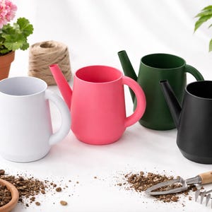 Op de afbeelding: Vier kleurrijke gieters in wit, roze, groen en zwart staan op een wit oppervlak. De gieters hebben een afgeronde vorm met een tuit en een handvat. Tuingereedschap en potplanten zijn ook in de afbeelding te zien.