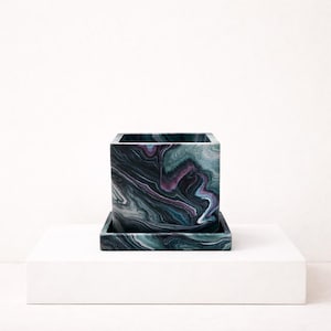 May include: Square planter with a matching tray. The planter has a marbled design in teal, purple, and black. The square shape and unique pattern make it a decorative piece for any home or office.