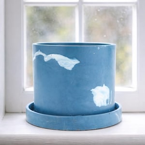 May include: A cylindrical blue planter with a matching saucer. The planter has white abstract paint details. The pot is sitting on a white windowsill in front of a window.